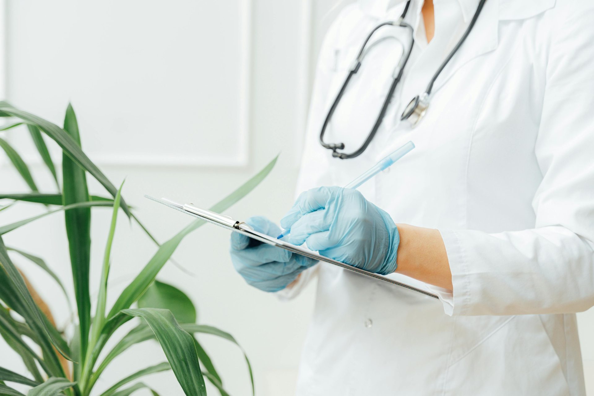 A healthcare professional wearing gloves takes notes on a clipboard with a stethoscope around their neck.
